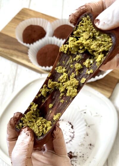 カカオパウダーがまぶされた外皮を割って、中の鮮やかな緑色のカダイフ（細い麺状の生地）を引っ張り出している様子。木製プレートの上には、カットされていないチョコトリュフが並んでいる。