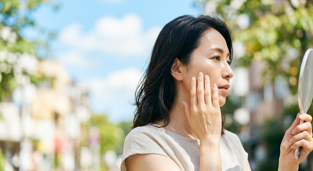 夏の強い日差しの下で手鏡を見ながら頬の毛穴やテカリを気にする40代50代の女性のイメージ