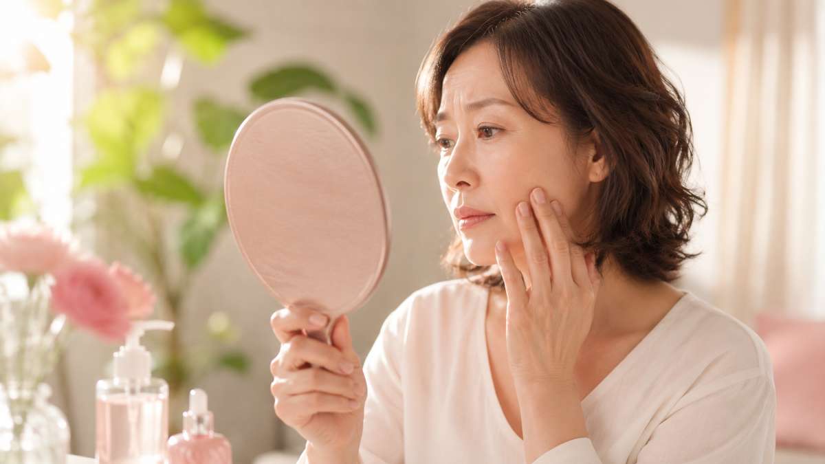 夏の終わりの窓辺で手鏡をのぞき込み、頬に指を添えてシミやくすみを気にする40代50代の女性のイメージ